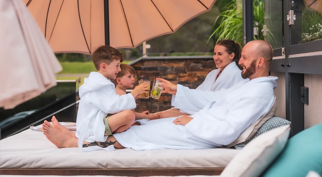 Eine Familie entspannt in Badem&auml;nteln unter einem Sonnenschirm und st&ouml;&szlig;t mit Getr&auml;nken an beim Familienurlaub im Hotel Kaprunerhof