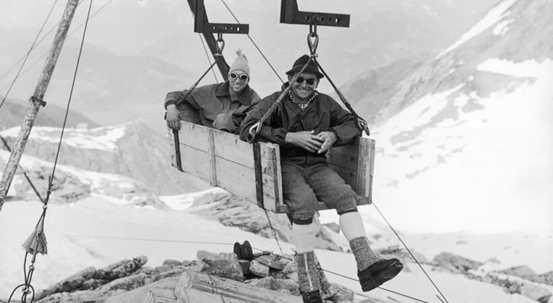 Zwei M&auml;nner sitzen in einer h&ouml;lzernen Seilbahn &uuml;ber verschneiten Bergen im Gletscherskigebiet am Kitzsteinhorn