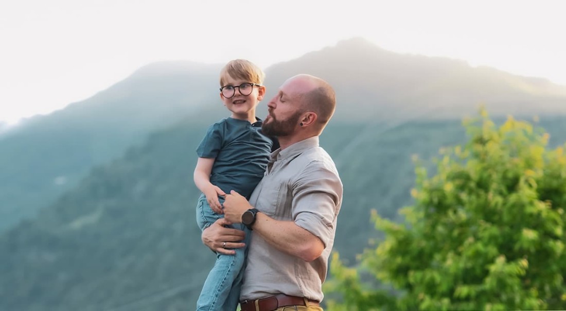 Mann h&auml;lt ein Kind im Freien vor einer Berglandschaft im Arm auf der Sonnenterrasse im Hotel Kaprunerhof