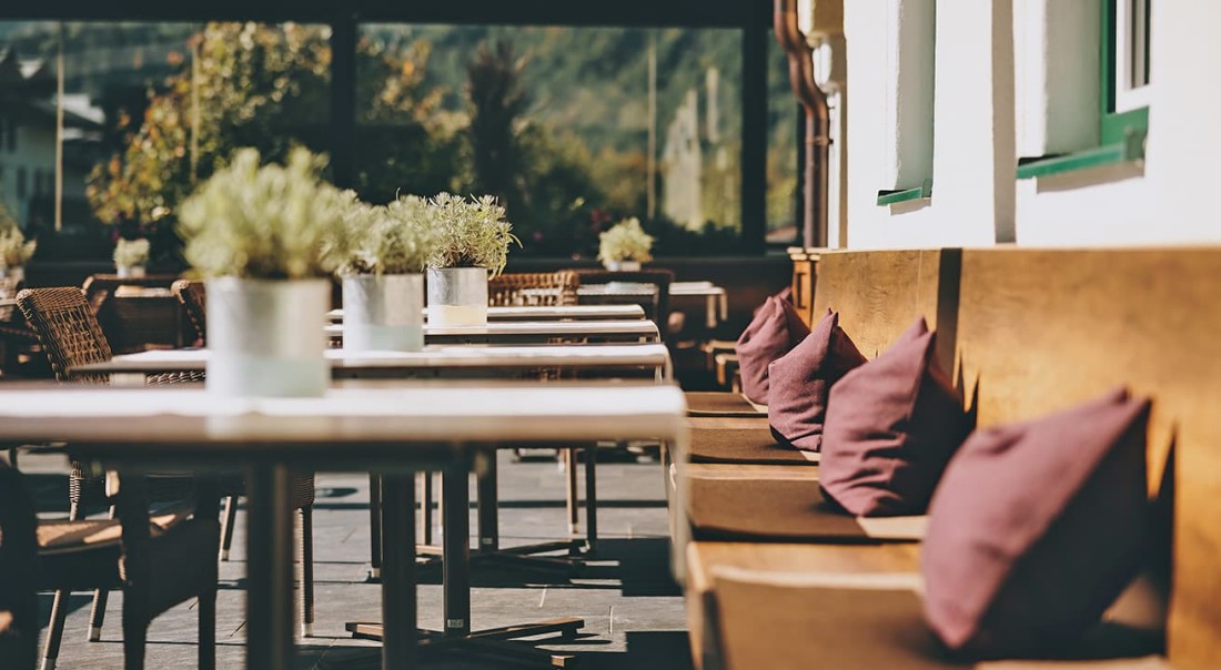 Sonnenbeschienene Terrasse mit Holzb&auml;nken, lila Kissen und Tischen mit Pflanzen im Hotel Kaprunerhof