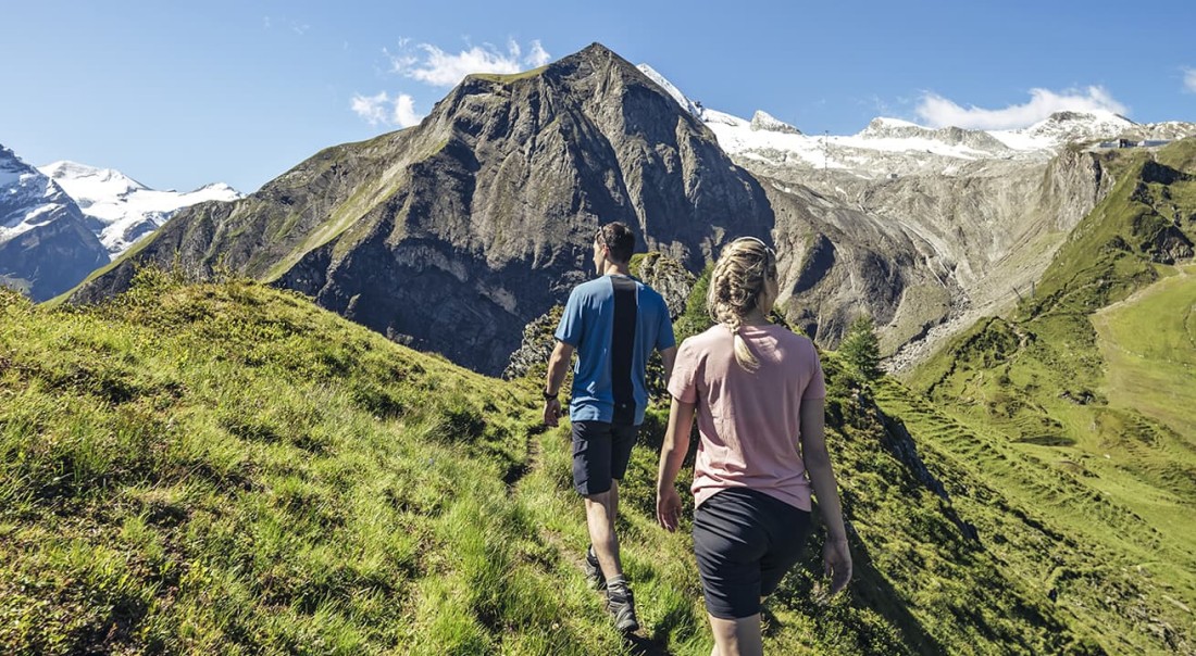 Zwei Menschen wandern auf einem gr&uuml;nen Bergpfad vor einer majest&auml;tischen Bergkulisse