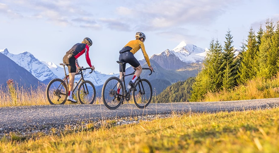 Zwei Radfahrer fahren auf einer Bergstra&szlig;e mit schneebedeckten Gipfeln im Hintergrund