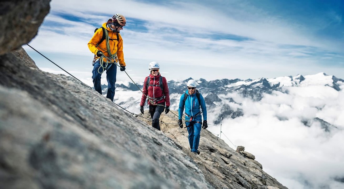 Drei Bergsteiger klettern an einem felsigen Hang am Kitzsteinhorn mit Schneegipfeln im Hintergrund