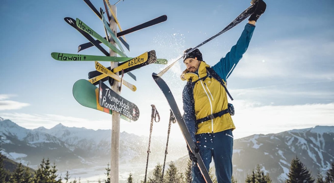 Ein Skifahrer hebt einen Ski in schneebedeckter Berglandschaft neben Wegweisern hoch